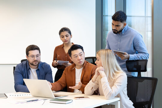 A diverse team collaborates around a table, discussing a laptop and sharing ideas. The focus is on teamwork and problem-solving.