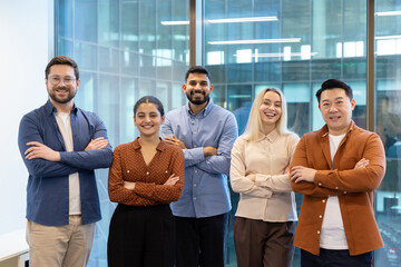 Group of diverse business professionals smiling and standing together indoors, arms crossed, near a...