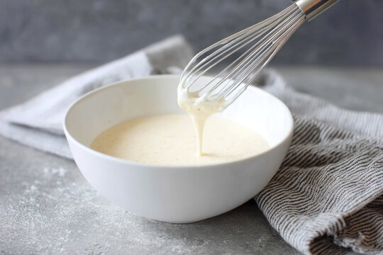Preparing creamy bechamel sauce in a white bowl with a whisk and kitchen towel on a textured surface