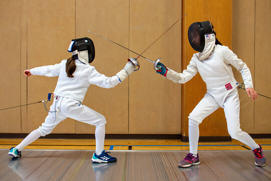 Two fencers in white gear lunge toward each other during an intense bout on an indoor fencing piste.