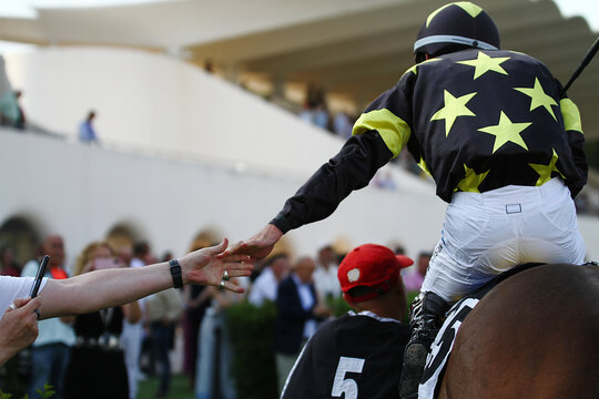Celebraci&oacute;n de un triunfo compartido dando la mano encima de un caballo entre jockey con chaquetilla de estrellas y aficionado de Turf en el hip&oacute;dromo