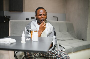 Portrait of a young African American man sitting on the couch at home and holding a throat spray in...