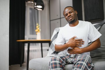 Man clutching his stomach and expressing discomfort while sitting on a sofa in a modern living room.