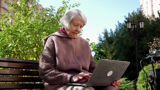 A stabilized image of an elderly modern woman with a laptop in the park on a bench. An octogenarian grandmother with a laptop in the courtyard of a multi-storey residential complex in autumn.