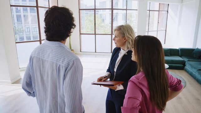 Woman in business attire presents a spacious apartment to a couple, highlighting large windows and urban views, showcasing potential living space