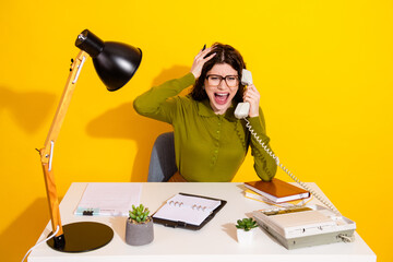Young woman expressing frustration during a phone call, sitting at a desk against a vibrant yellow...