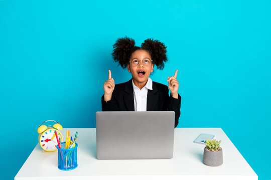 Excited schoolgirl at desk with laptop, vibrant teal background, expressing enthusiasm for learning and creativity