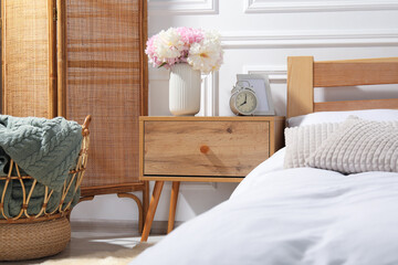 Bedside table with alarm clock and flowers near bed indoors