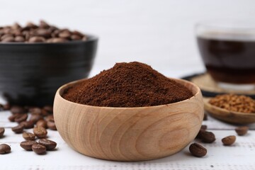 Instant coffee powder in bowl and beans on white wooden table, closeup