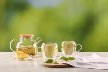 Tasty green tea in glass cups, teapot, lemon and leaves on white marble table outdoors