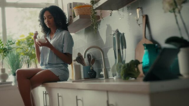  a woman with curly hair sitting on a kitchen counter near a window, holding a bowl and smiling while eating. She is wearing a striped shirt, and there are various potted plants and kitchen.