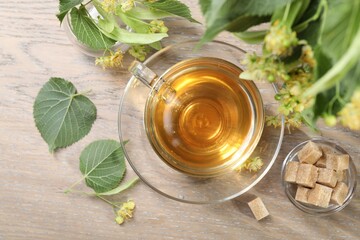 Tasty linden tea in cup, sugar cubes, leaves and flowers on wooden table, flat lay