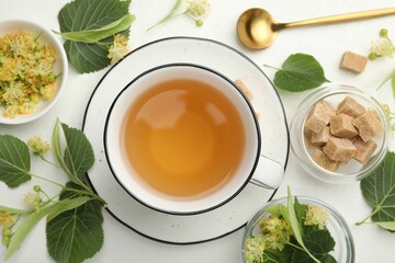 Tasty linden tea in cup, spoon, sugar cubes, leaves and flowers on white table, flat lay