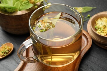 Tasty linden tea in cup, leaves and flowers on dark textured table, closeup