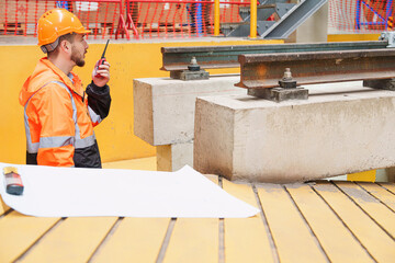 Engineer in orange safety gear uses walkie-talkie beside train rail in modern maintenance facility....