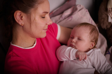 Young adult woman with brown hair cradles sleeping baby girl. Calm expression. Cozy room with warm...
