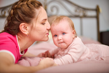 Young mother with braided hair kisses light-haired baby girl on pink bedspread, expressing warmth and love. Soft lighting in cozy bedroom setting enhances tenderness