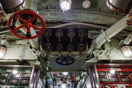 The cluttered and complex interior of a vintage submarine, showing a control panel with multiple pressure gauges, large red and black valve wheels, and a dense network of pipes and wiring on the ceili