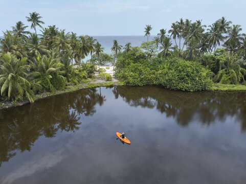Aerial view of a lone figure paddling an orange kayak across the still, dark waters of a lagoon, framed by lush greenery, Kamadhoo, Baa Atoll, Maldives.