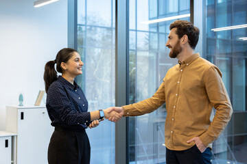 Two smiling business people shake hands in a modern office setting, likely sealing a deal or...