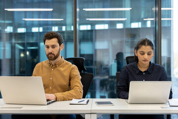 Two diverse colleagues work diligently at their laptops in a modern office, focused on their tasks.