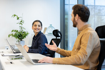 In an office, a man and a woman are seated at a desk, collaborating on a project while using...