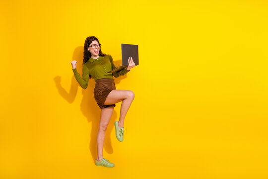 Happy young woman celebrating success holding laptop on a vibrant yellow background wearing stylish green and brown outfit