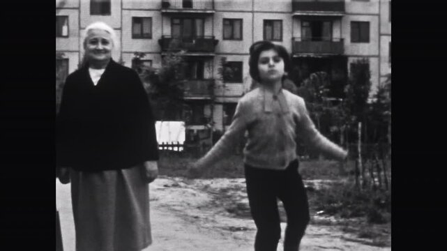 Young girl active jump rope while elderly women stand nearby smiling. Child joy contrasted with calm elder presence in outdoors. Vintage film. Old archive video. Archival retro 1960s happy childhood