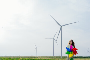 Fototapeta premium Young woman with long hair in linen trousers and green shirt holding colorful pinwheel on open field. Wind turbines in distance. Concept of visibility, protest and climate optimism.