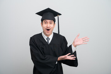 A young man in a graduation gown and cap smiles at the camera, striking a confident pose to...