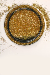 A black circular object is filled with shimmering gold glitter, with some glitter scattered around it on a clean white surface, creating a festive composition