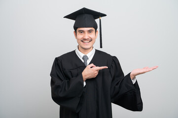 A young man in a graduation gown and cap smiles at the camera, striking a confident pose to...