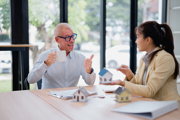 Real estate agent presenting a house model to a cheerful customer enjoying coffee and giving a thumbs-up during an engaging meeting in a modern office setting