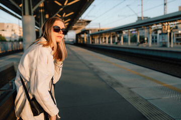 Fototapeta premium Stylish blonde woman with sunglasses and shoulder bag is waiting for train at sunset, sitting on bench in railway station, enjoying golden hour light. Woman leaning to see if train is coming