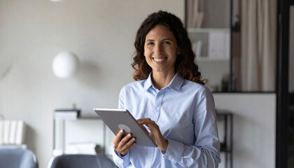 Portrait of young Hispanic professional business woman standing in office. Happy female company executive, smiling businesswoman entrepreneur corporate leader manager looking at camera using tablet.
