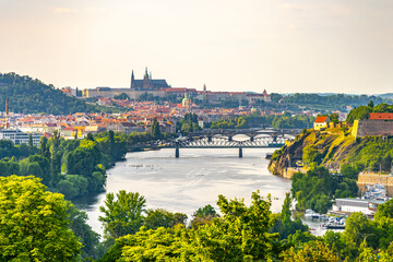Obraz premium A breathtaking view of Prague showcasing the Vltava River winding through the city. The historic skyline features iconic landmarks, surrounded by lush greenery, creating a serene atmosphere.
