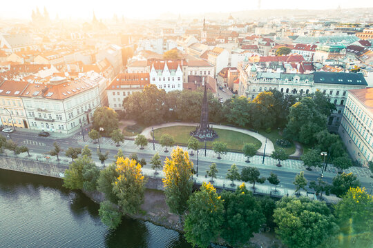 Visitors admire Kranner's Fountain surrounded by lush greenery in the Park of the National Awakening, overlooking the Vltava River and historic buildings of Prague during sunset.