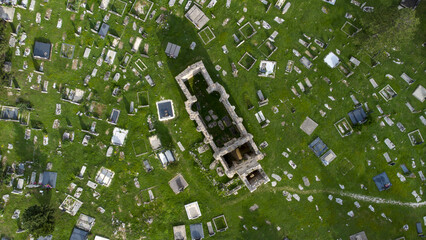 Aerial view of a graveyard with scattered, aged headstones among lush grass and a roofless stone structure, [Actual City Name], [Actual State Name], [Actual Country Name].