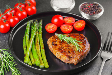 Tasty grilled beef steak served with green asparagus and cherry tomatoes on black plate, with cutlery. Perfect for food menu, diet, or culinary design concepts.