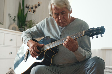 Senior man is playing guitar. Elderly male sitting at sofa and play music. Portrait of a gray...