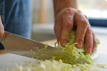 Handsome bearded man is preparing green fresh vegan salad in the kitchen at home. Male cutting vegetables, lettuce with knife on wooden chopping board, cooking vegetarian meal. Tasty healthy diet food