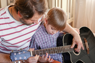 Handsome dad teaches his cute son to play the guitar sitting on sofa in the living room at home....