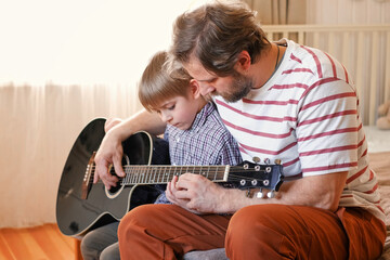 Handsome dad teaches his cute son to play the guitar sitting on sofa in the living room at home....