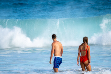 Couple standing on sandy beach, gazing at a powerful wave crashing under a moody sky. Moment of awe, connection, and raw nature. Adventure captured by the ocean’s edge.