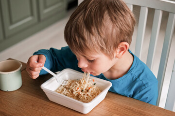 6 years old boy eats instant noodle from box. Child is eating spaghetti. Lunch time. A good appetite. Tasty children food. Close-up portrait. Carbs dinner. Fast food. Kid eating by himself with fork.