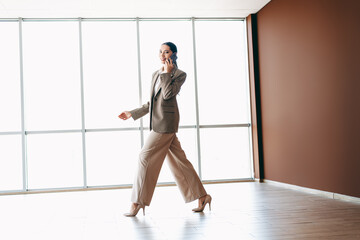 Professional businesswoman walking indoors holding a phone, dressed in an elegant formal suit,...