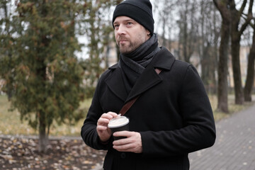 Pensive hipster drinking coffee in the urban street. Handsome man wearing black coat, brown leather...