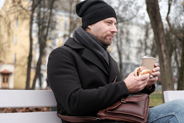 Pensive hipster drinking coffee sitting on a bench in the urban park. Handsome man wearing black coat eating street food, hot dog with paper cup in hand in the city. Stylish bearded middle-aged male.
