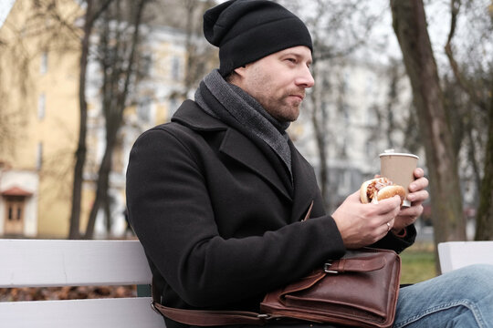 Pensive hipster drinking coffee sitting on a bench in the urban park. Handsome man wearing black coat eating street food, hot dog with paper cup in hand in the city. Stylish bearded middle-aged male.