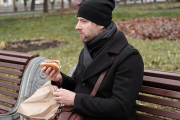 Pensive hipster drinking coffee sitting on a bench in the urban park. Handsome man wearing black...